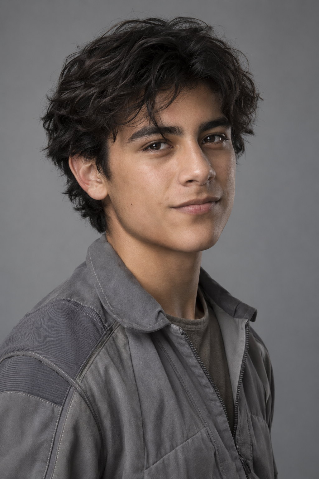 Santi, a teenager with dark curly hair, in a grey flight suit with the collar open, half-smiling at the camera.